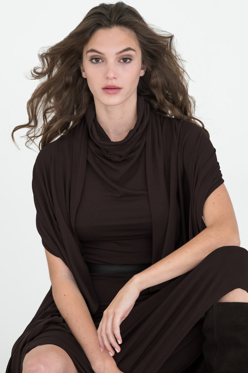 A woman with long, wavy brown hair wearing a white sleeveless top with a high draped collar, photographed in a bright indoor setting near a window.