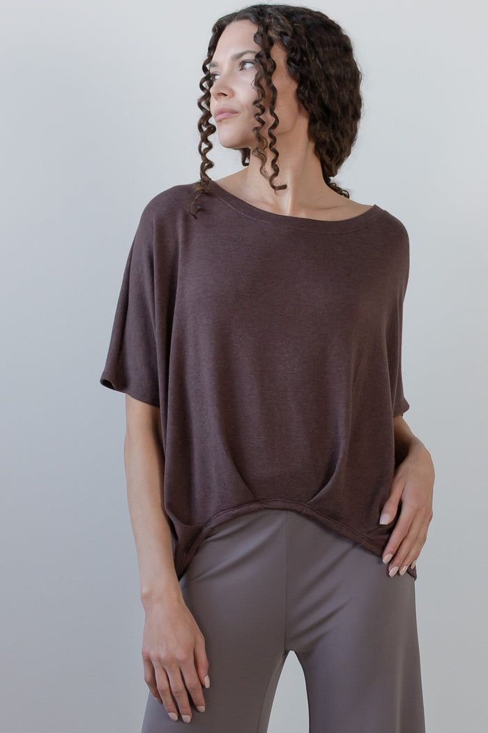 A woman wearing a loose, short-sleeve scoop-neck top in a muted dark brown or taupe-brown color, paired with gray leggings, photographed against a plain light gray studio background.