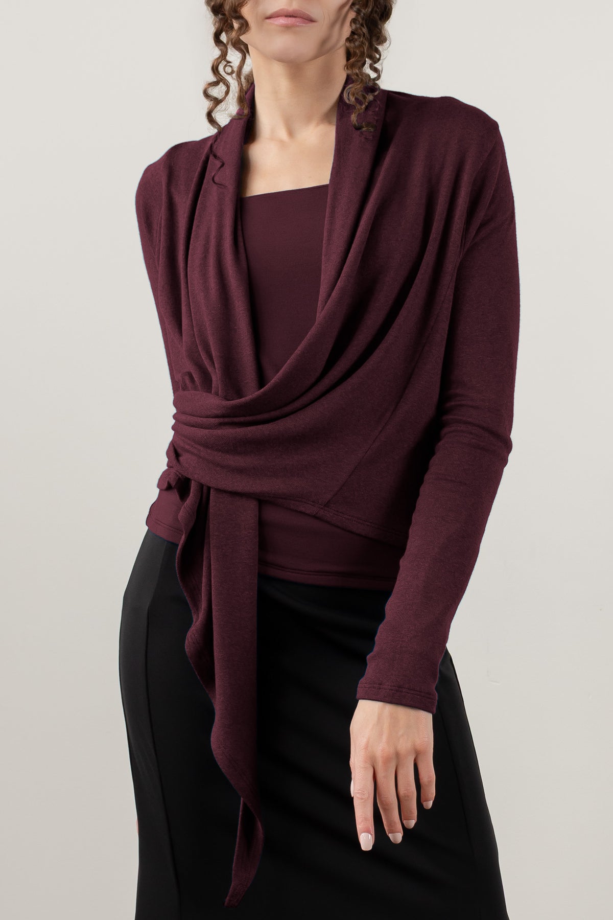 A woman wearing a dark burgundy wrap cardigan with a cowl neck and tie detail, styled over a matching top and black skirt against a light gray studio background.