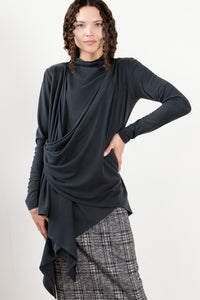 A model wearing a black long-sleeve turtleneck top, a black draped open-front cardigan, a gray and black plaid skirt, black tights, and black boots, standing against a plain off-white studio wall.