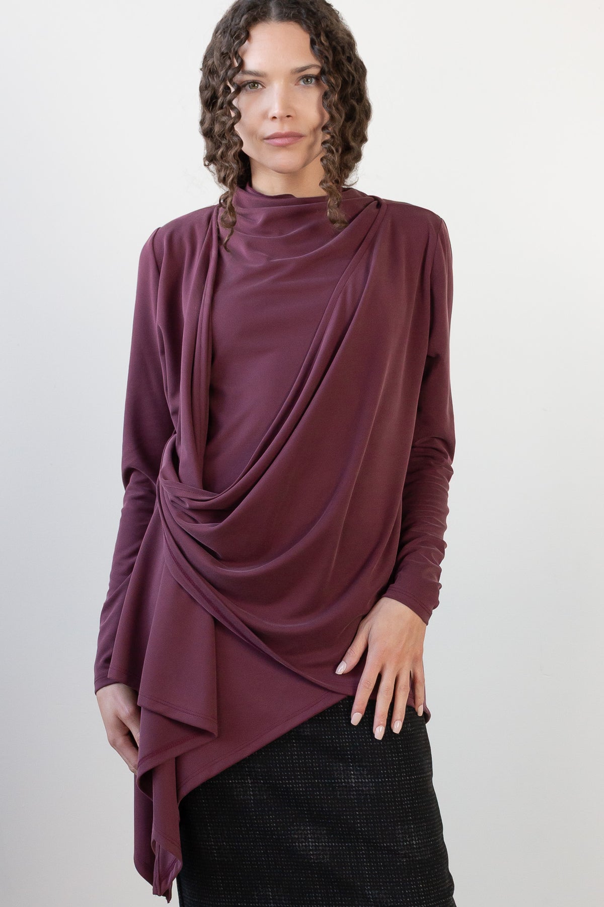 A model wearing a draped, long-sleeve top in a deep plum purple color, paired with a black plaid skirt and black boots, standing against a plain white studio background.
