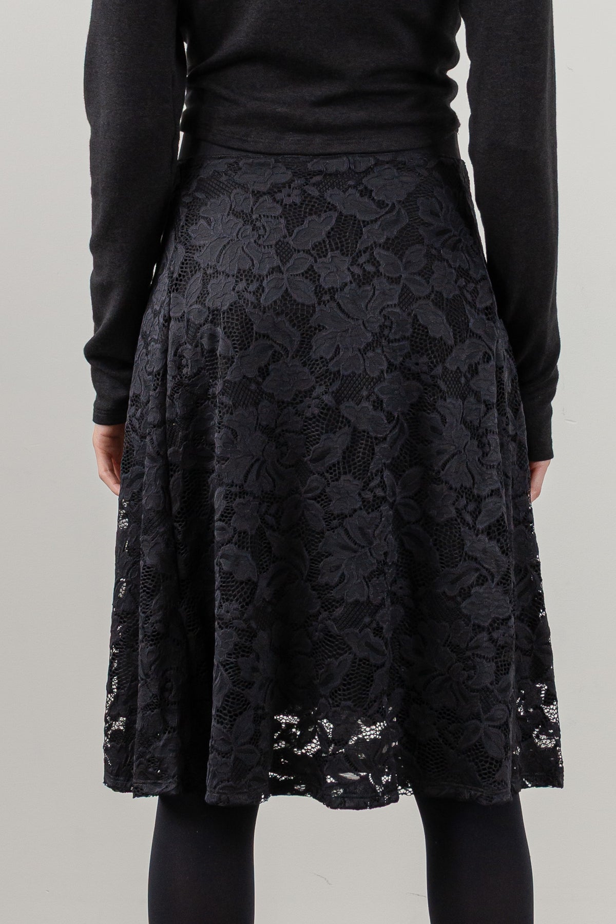 A black lace A-line skirt with a floral pattern, worn by a model in a black long-sleeve top and black tights against a light gray studio background.