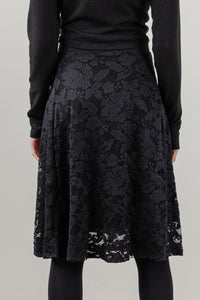 A black lace A-line skirt with a floral pattern, worn by a model in a black long-sleeve top and black tights against a light gray studio background.