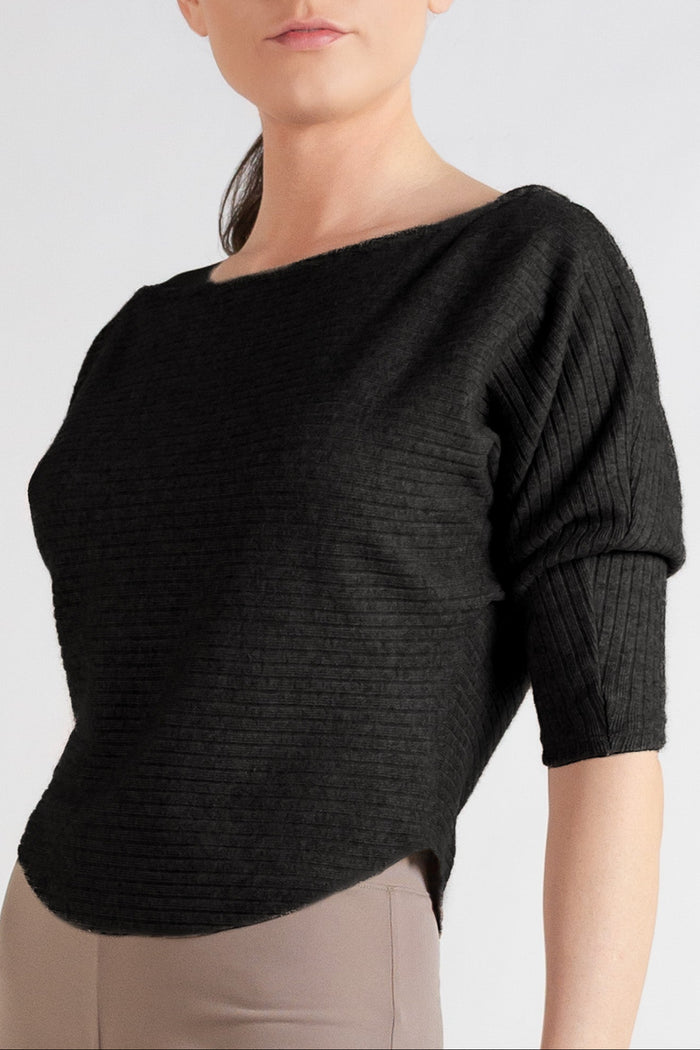 A woman modeling a black off-the-shoulder three-quarter sleeve top and taupe wide-leg pants against a light gray studio background.