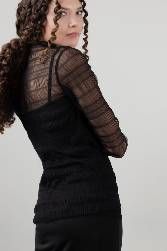 A model wearing a black sheer mesh long-sleeve top and matching pleated mini skirt with a wide waistband, photographed against a plain light gray studio background. Back view.