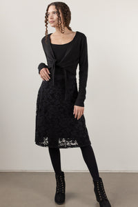 A woman modeling a black long-sleeve wrap cardigan tied at the waist over a black scoop-neck top, paired with a black lace midi skirt, black tights, and black lace-up ankle boots, against a plain light beige studio background.