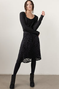 A woman modeling a black long-sleeve knit cardigan over a black top and a black lace midi skirt, styled with black tights and black lace-up ankle boots against a plain light gray studio background.