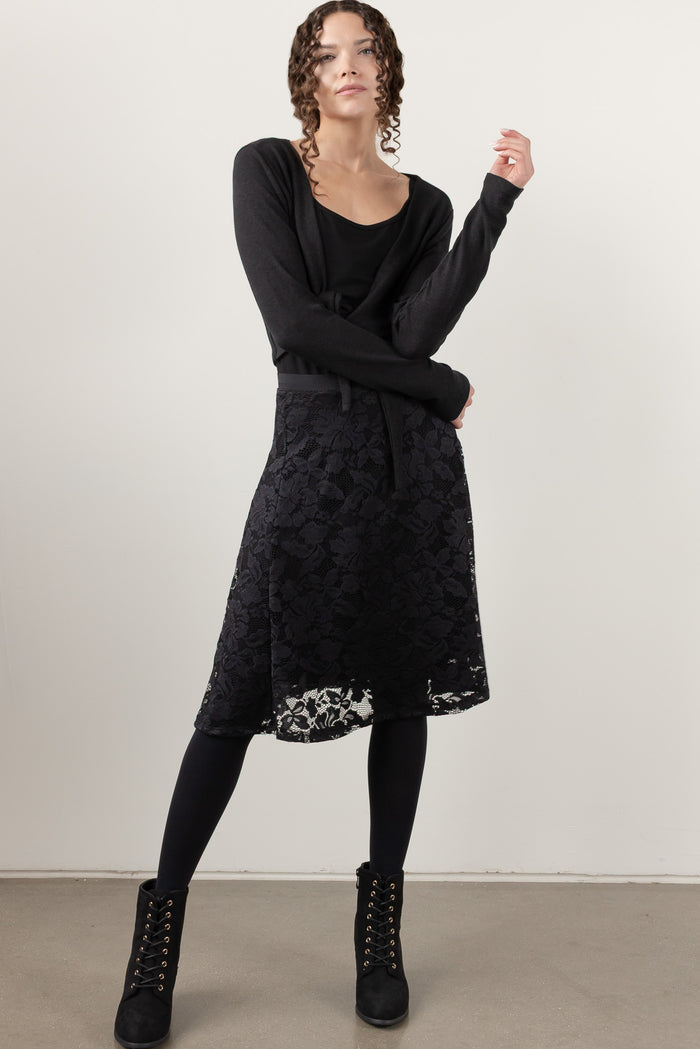A woman modeling a black long-sleeve knit cardigan over a black top and a black lace midi skirt, styled with black tights and black lace-up ankle boots against a plain light gray studio background.