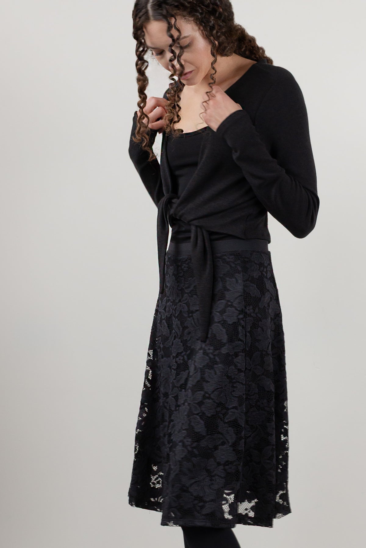 A woman modeling a black long-sleeve knit wrap top and a black lace midi skirt against a plain light gray studio background.
