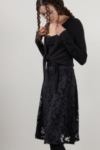 A woman modeling a black long-sleeve knit wrap top and a black lace midi skirt against a plain light gray studio background.
