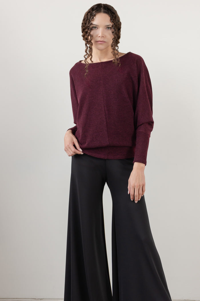 A woman wearing a loose, boat-neck knit sweater in deep burgundy with black pants, shown from the nose down against a light gray studio background.