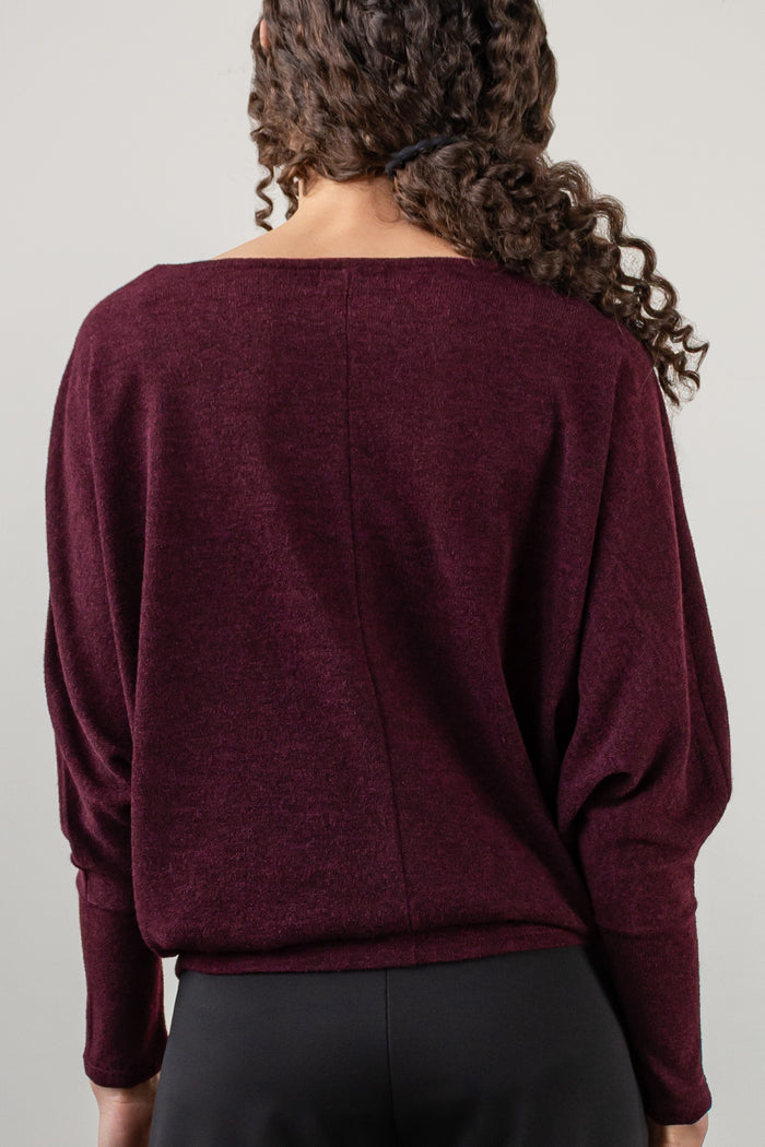 A woman wearing a loose, boat-neck knit sweater in deep burgundy with black pants, shown from the nose down against a light gray studio background. Back view.