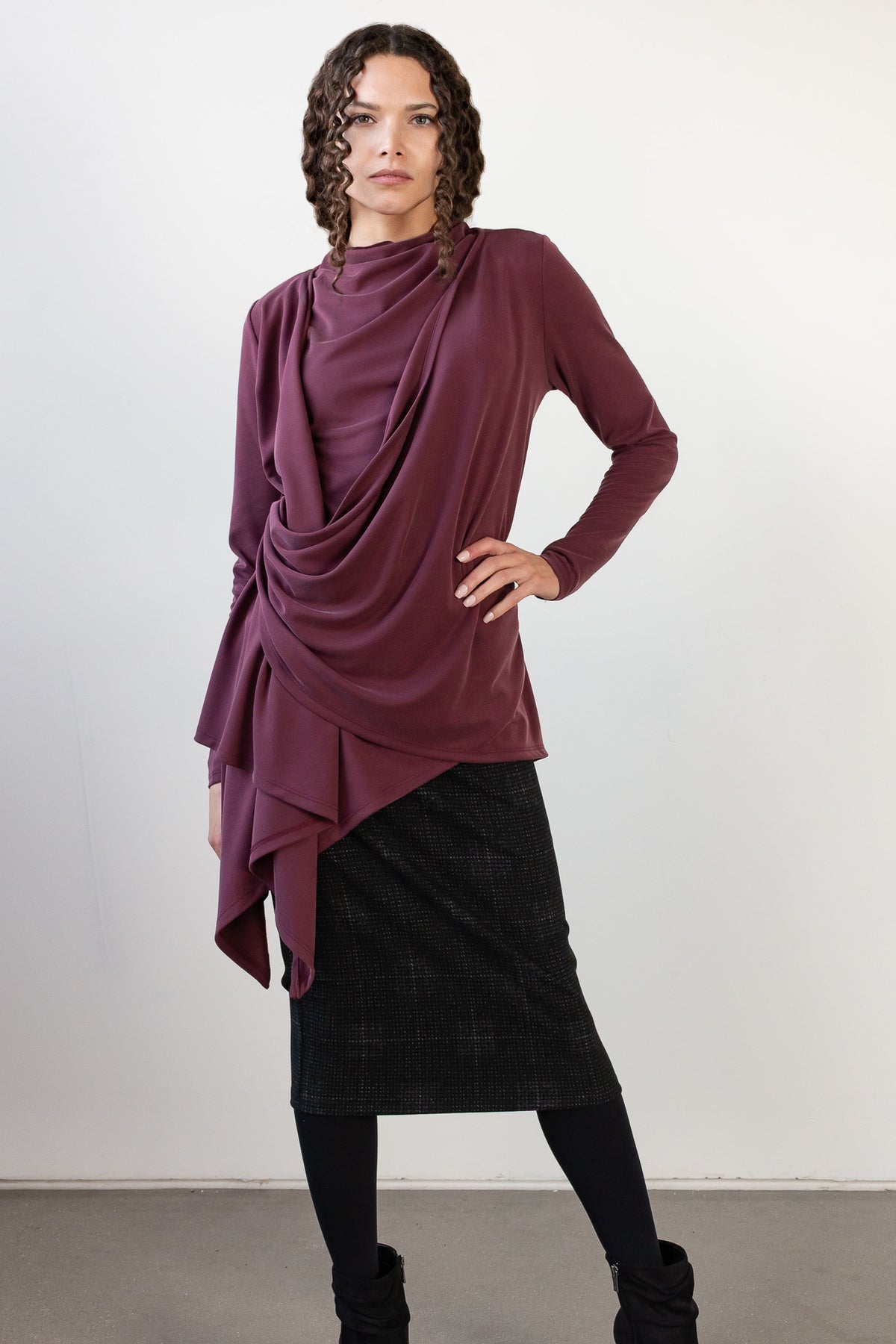 A model wearing a draped, long-sleeve top in a deep plum purple color, paired with a black plaid skirt and black boots, standing against a plain white studio background.