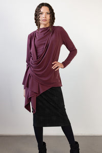 A model wearing a draped, long-sleeve top in a deep plum purple color, paired with a black plaid skirt and black boots, standing against a plain white studio background.