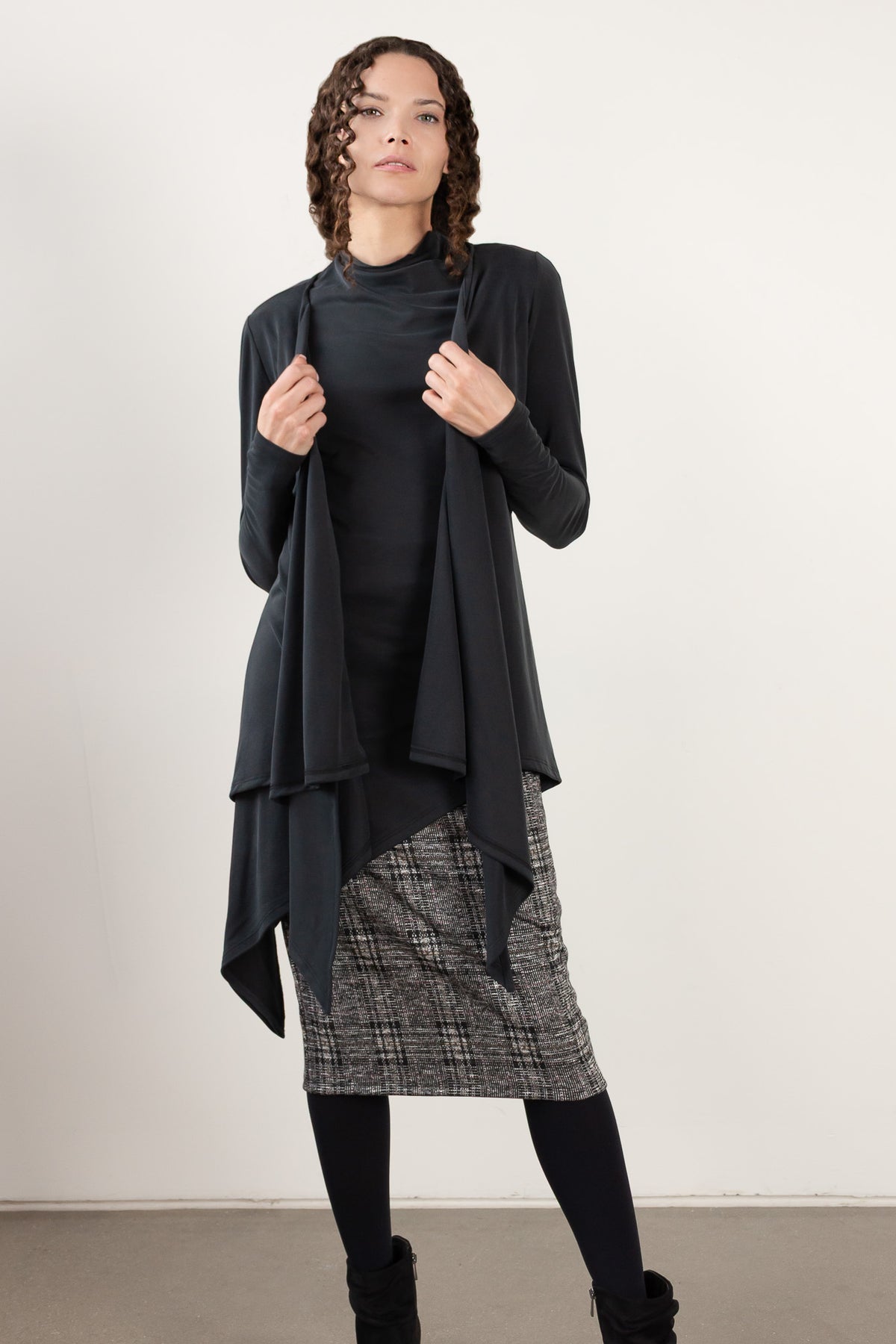A model wearing a black long-sleeve turtleneck top, a black draped open-front cardigan, a gray and black plaid skirt, black tights, and black boots, standing against a plain off-white studio wall.