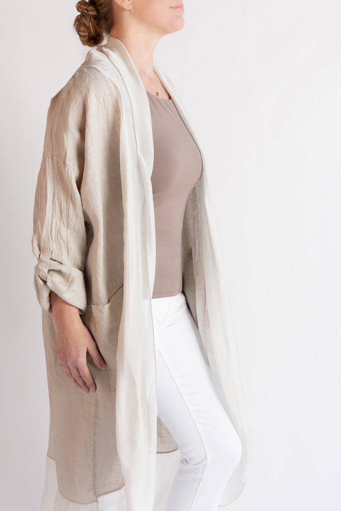 Woman wearing a beige long coat over a brown top and white pants on a plain background