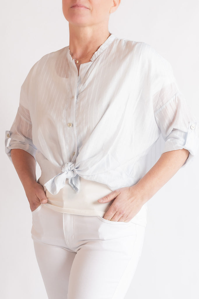 Person wearing a light-colored shirt with rolled-up sleeves and tied waist, standing against a white background.