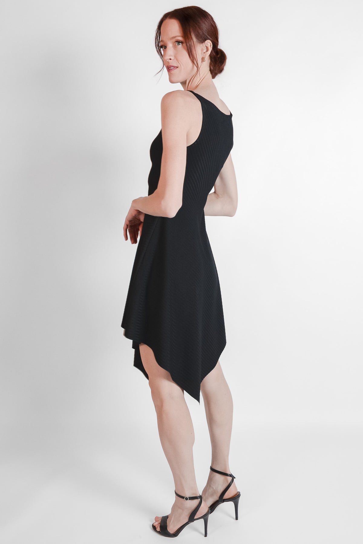 Woman wearing black ribbed fabric dress with an asymmetrical hem. Side view.