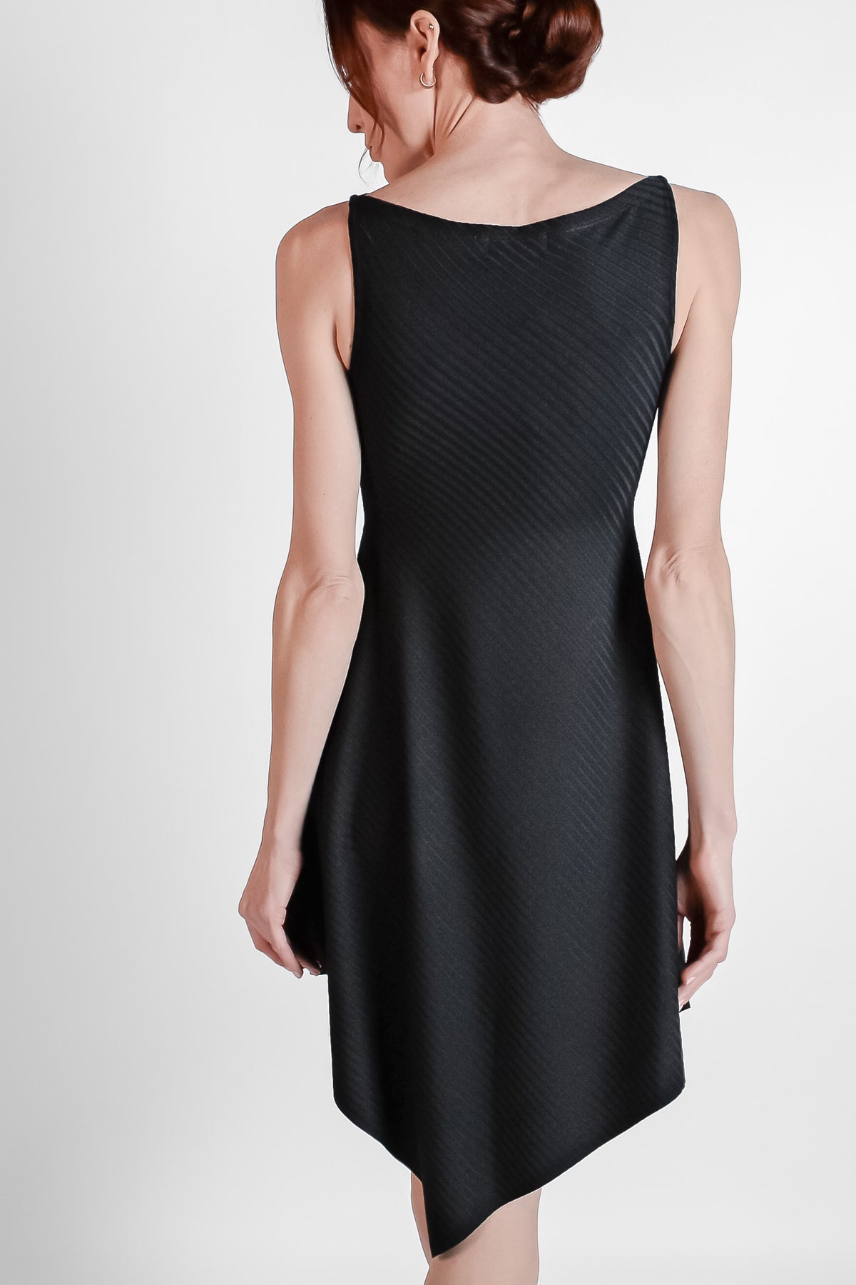 Woman wearing black ribbed fabric dress with an asymmetrical hem. Back view.