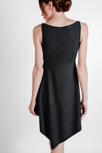 Woman wearing black ribbed fabric dress with an asymmetrical hem. Back view.