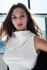 A woman with long, wavy brown hair wearing a white sleeveless top with a high draped collar, photographed in a bright indoor setting near a window.