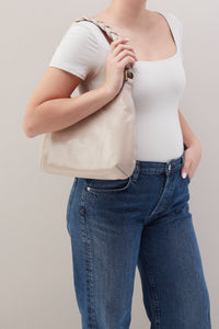 Person wearing a white top and blue jeans, holding a beige handbag against a plain background