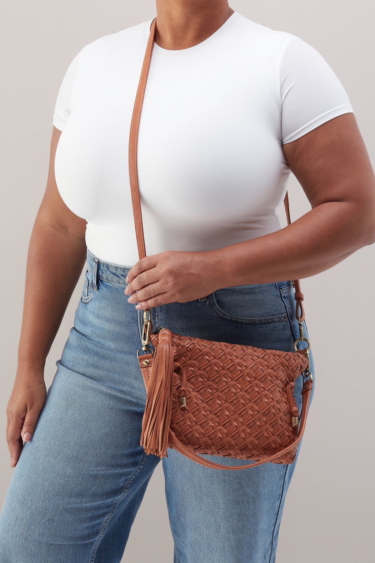 Person wearing a white top and blue jeans holding a brown woven handbag against a neutral background