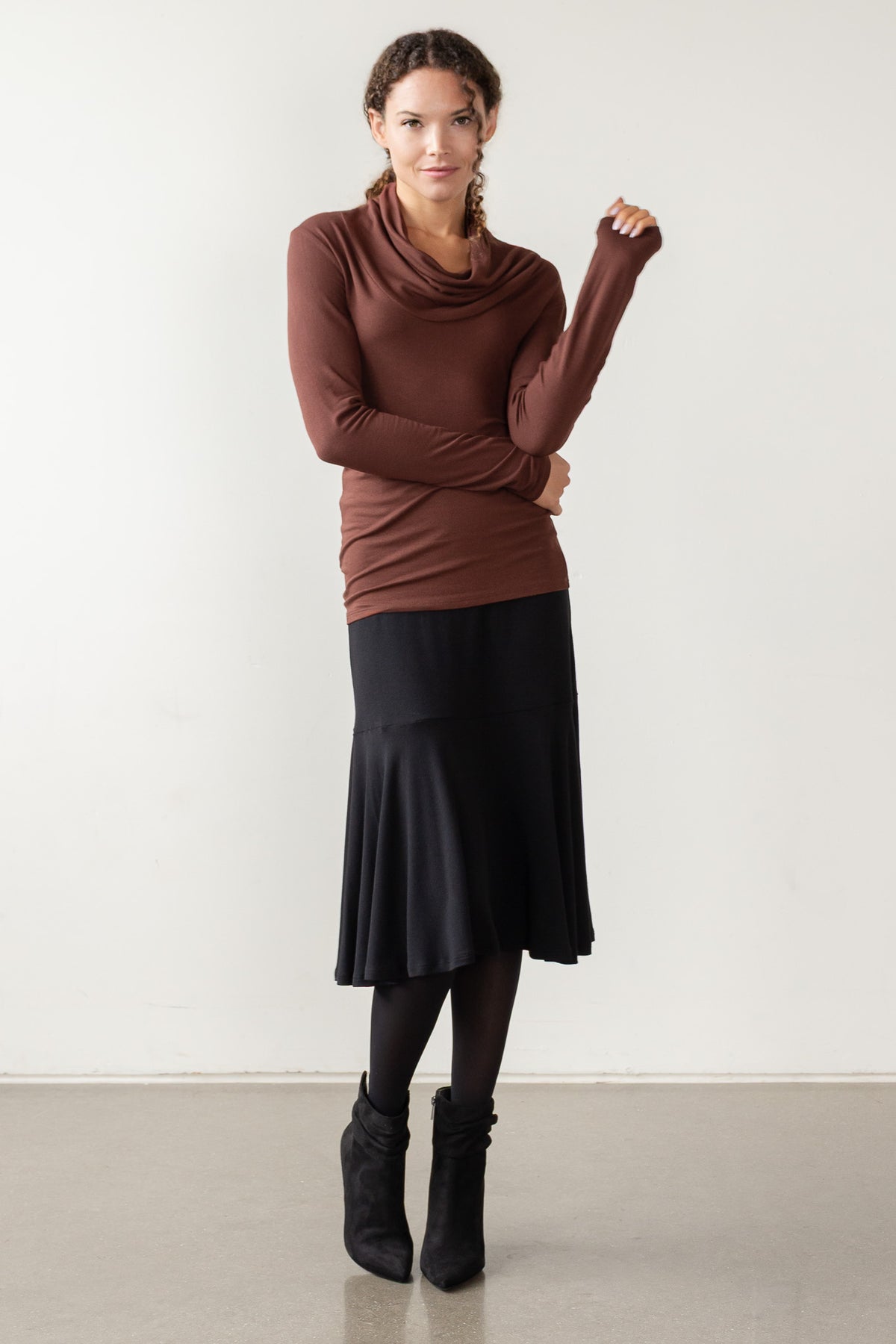 Woman wearing a brown long-sleeve top with a cowl neck and black skirt against a white background.